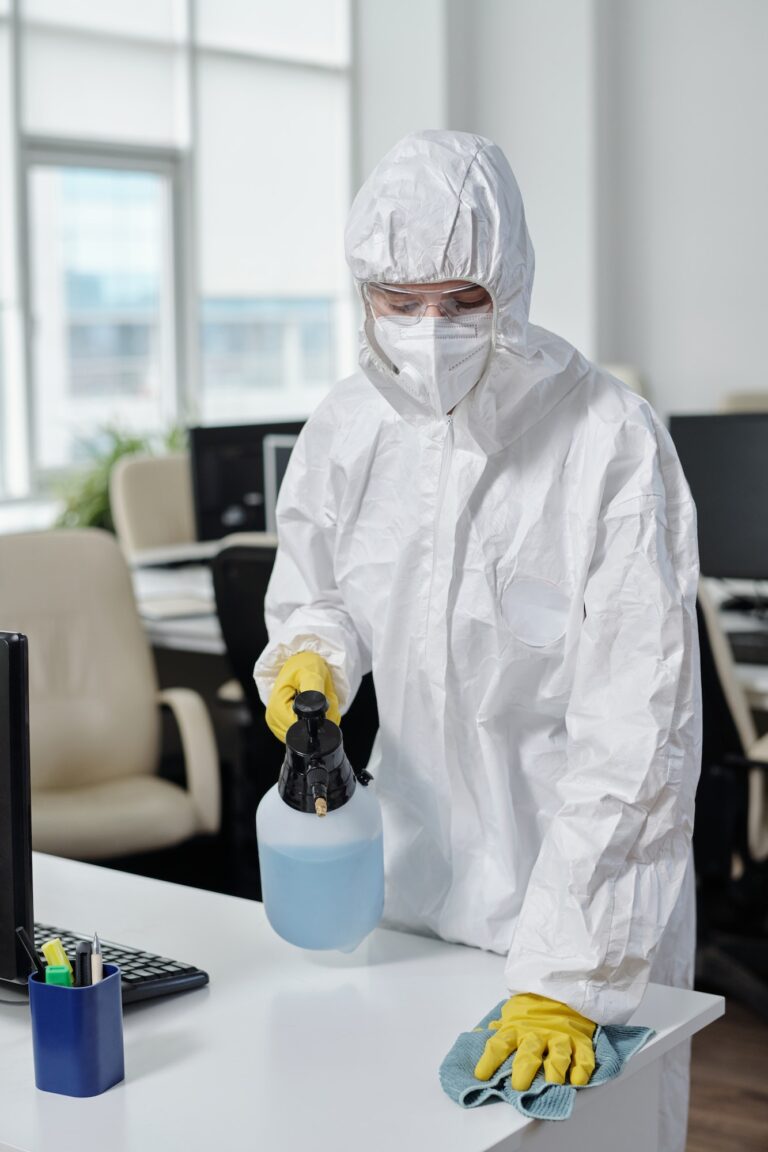 Contemporary cleaning service worker with sprayer disinfecting desks in office